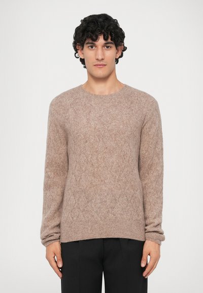 Homme aux cheveux bouclés et foncés portant un pull en tricot beige texturé et un pantalon noir, se tenant devant un fond clair uni.