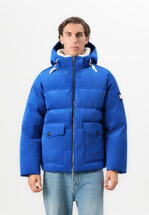 Man wearing a blue corduroy puffer jacket with hood and front flap pockets, standing against a plain white background.
