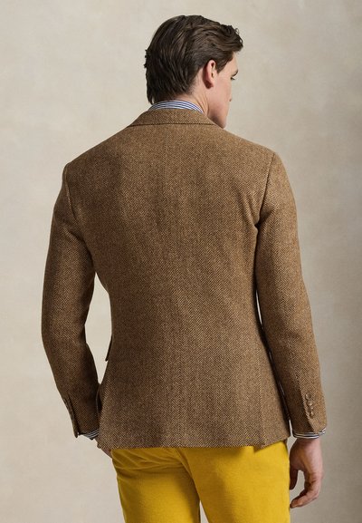 Άνδρας γυρισμένος με καφέ blazer ψαροκόκκαλο, ριγέ πουκάμισο και φωτεινό κίτρινο παντελόνι σε απλό μπεζ φόντο.