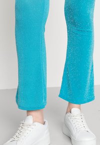 Leggings turquesa com uma textura cintilante e bainha larga, combinados com ténis brancos com acabamento suave e design de atacadores.
