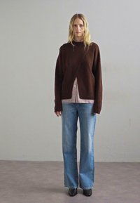 Καφέ πλεκτό πουλόβερ με ασύμμετρο σχέδιο, επενδυμένο με μια ριγέ μπλούζα, συνδυασμένο με ανοιχτό μπλε τζιν παντελόνι φαρδιάς γραμμής και μαύρες γόβες.