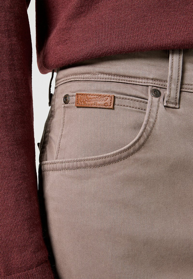 Jeans beige con una texture liscia, caratterizzati da un'etichetta in pelle, una tasca frontale e dettagli di cucitura a contrasto. Abbinati a un maglione bordeaux.