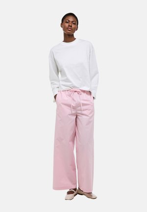 Femme debout, les mains dans les poches, portant une chemise blanche à manches longues, un pantalon rose ample avec un cordon de serrage, et des chaussures plates beiges.