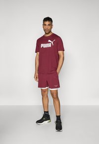 T-shirt de sport maroon avec logo "PUMA", shorts assortis et baskets noires avec des accents verts. Présente un design simple et fonctionnel.