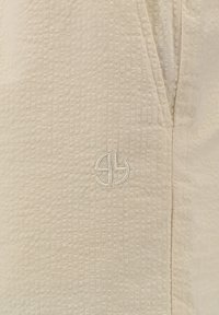 Close-up van een beige gestructureerde stof met een klein, geborduurd rond logo nabij een verticale naad.