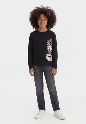 Enfant aux cheveux bouclés debout, portant un t-shirt noir à manches longues avec un texte coloré vertical et un jean noir délavé avec des baskets blanches.