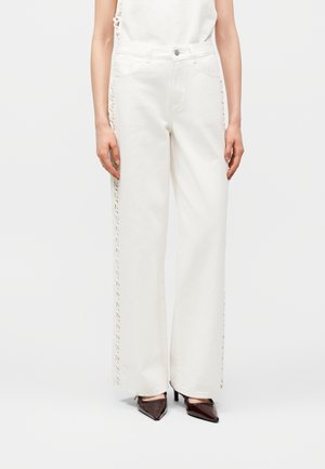 Femme portant un pantalon blanc taille haute à jambes larges avec des détails à lacets sur les côtés et des talons pointus foncés, debout devant un fond uni.