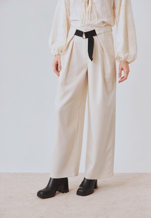 Mujer vistiendo pantalones anchos color crema con un cinturón negro y una blusa blanca de manga larga bordada, combinados con botines negros de tacón.