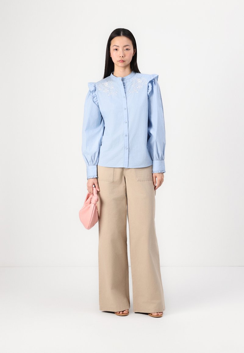 Chemise bleu clair à manches froncées et détails brodés, associée à un pantalon large beige et un sac à main rose.