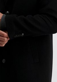 Zwarte wolcoat met een knoopmanchet, met een grijze gebreide textuur bij de mouw. Zichtbare knopen langs de manchet voegen detail toe.