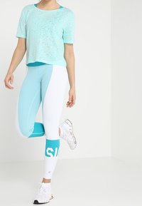 Ljus teal kortärmad tröja med textur, i kombination med vita och teal leggings med färgblocking och logotyp. Vita träningsskor.
