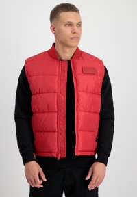 Gilet rembourré rouge avec fermeture éclair à l'avant et col côtelé. Présente une texture matelassée et une petite étiquette logo sur la poitrine. Porté par-dessus un haut noir.