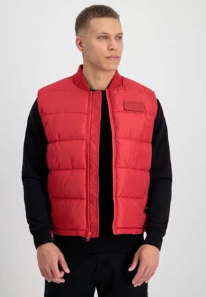 Rode puffer vest met een ritssluiting aan de voorkant en een geribde kraag. Heeft een geborduurd patroon en een klein logo op de borst. Draagt over een zwart top.