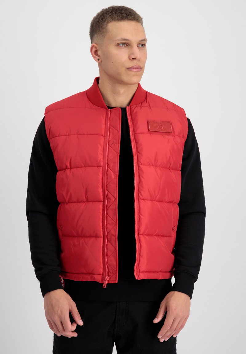 Gilet rembourré rouge avec fermeture éclair à l'avant et col côtelé. Présente une texture matelassée et une petite étiquette logo sur la poitrine. Porté par-dessus un haut noir.