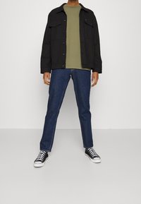 Černá bunda přes olivově zelenou košili, doplněná modrými džíny a černými teniskami. Ležérní, volný střih s knoflíkovým zapínáním.