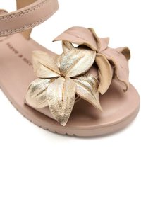 Sandalia color marrón con una tira texturizada de color rosa claro y un gran acento de flor metálica dorada en la parte frontal. Diseño de suela plana.