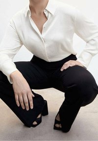 Personne portant une chemise blanche à boutons, un pantalon noir à jambes larges et des chaussures à talons ouvertes noires, accroupie les mains reposant sur les genoux.