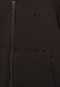 Tmavě hnědá bunda na zip s předními kapsami a jemně vyšitou značkou na levé straně hrudi.