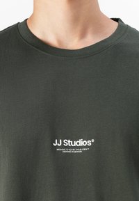 Olive green T-shirt with a crew neck, made of cotton, featuring white text "JJ Studios®" and additional branding beneath. Smooth texture.