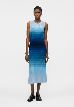 Γυναίκα που στέκεται, φοράει μίντι φόρεμα χωρίς μανίκια με ριμπ ύφανση και μπλε διαβαθμισμένες ρίγες, καθώς και μαύρα μοκασίνια, μπροστά σε απλό λευκό φόντο.