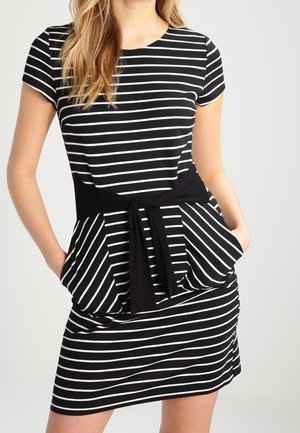 Vrouw in een zwart kort mouwen jurkje met witte horizontale en diagonale strepen, een zwarte tailleband en met haar handen in de zakken.