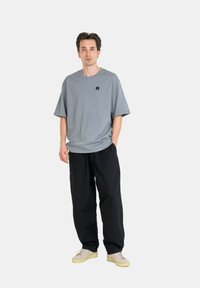 Graues T-Shirt mit kurzen Ärmeln und einem kleinen schwarzen Logo auf der Brust, kombiniert mit locker sitzenden schwarzen Hosen und hellen Sneakers.