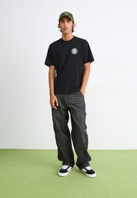 Schwarzes T-Shirt mit kurzen Ärmeln und einem kreisförmigen Logo auf der Brust, kombiniert mit dunkelgrauen weiten Hosen und schwarzen Schuhen mit weißen Sohlen.