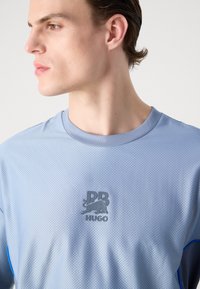 Light blue short-sleeve athletic shirt with a textured pattern and grey logo on the chest. Features a ribbed crew neckline and blue accents.
