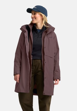 Woman wearing a mauve long raincoat, dark pullover, olive pants, and navy cap, smiling and looking to the right against a plain background.