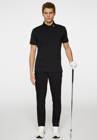 Svart pikétröja med tre knappar och en logotyp på kragen, tillsammans med svarta byxor och vita golfsko, håller i en golfklubba.