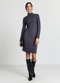 Eng anliegendes, langärmliges graues Kleid mit hohem Kragen; verfügt über eine glatte, dehnbare Textur. Kombiniert mit kniehohen schwarzen Stiefeln und einer kleinen Tasche.