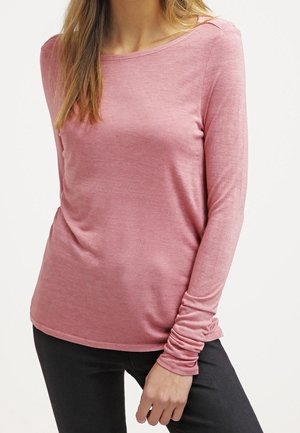 Femme portant une chemise rose à manches longues et un pantalon noir, debout avec une main posée près de sa hanche sur un fond uni.