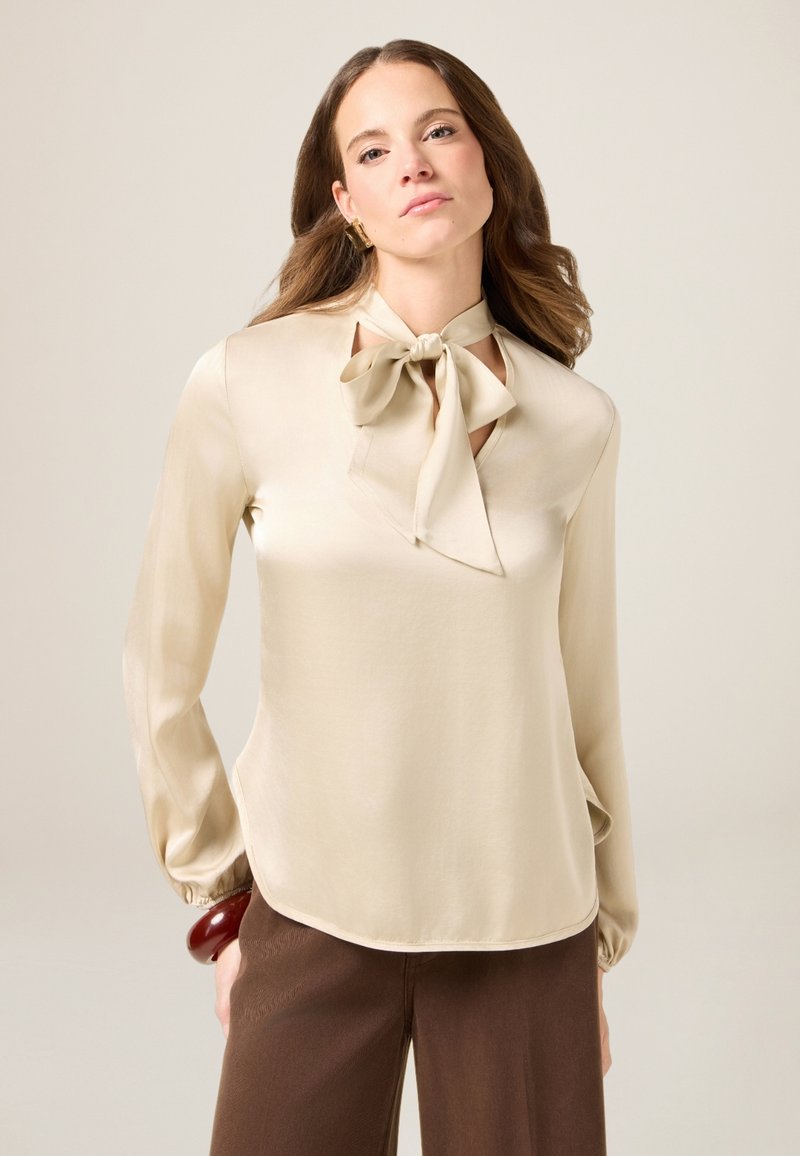 Beige Satinbluse mit einer Schleife am Kragen, langen Ärmeln mit gerafften Bünden und einer leichten A-Linie. Glatte Textur und elegantes Design.