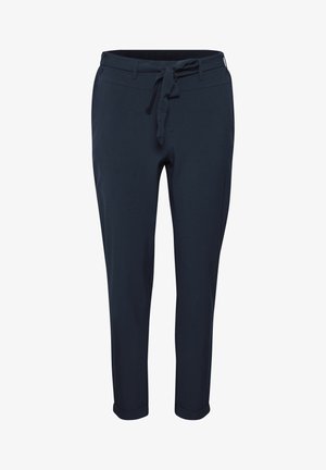 Femme debout, jambes croisées, portant un col roulé blanc, un pantalon bleu marine ceinturé et des talons noirs sur un fond clair uni.