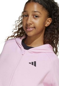 Sudadera con cremallera de color rosa claro con un acento de logo negro en la parte superior izquierda. La tela parece suave y tiene un corte relajado y capucha.