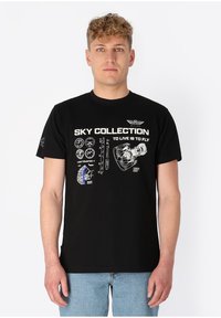 Mann trägt ein schwarzes T-Shirt mit weißen technischen Luftfahrtgrafiken und dem Text "Sky Collection To Live Is To Fly", kombiniert mit hellblauen Jeans.
