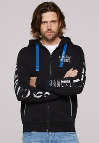 Sudadera negra con cremallera, cuerdas azules, texto gráfico blanco en las mangas y en la parte delantera. Tela suave, bolsillos tipo canguro, puños y dobladillo acanalados.