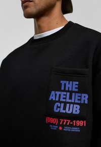 Zwarte sweatshirt met een voorkant zak. Blauwe tekst leest "THE ATELIER CLUB" met een rood telefoonnummer en aanvullende details eronder.