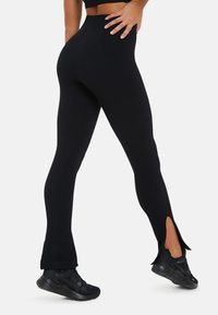 Zwarte geribde legging met een hoge taille, uitlopende onderkant en zijsplitten. Gecombineerd met zwarte sportschoenen voor een complete look.