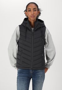 Veste sans manches noire zippée avec capuche, présentant un matelassage en chevrons, des cordons de serrage réglables et des poches latérales. Portée sur un sweatshirt gris.