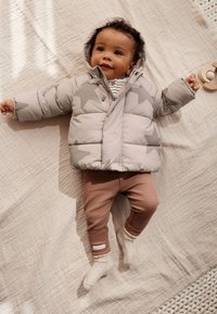 Veste matelassée à capuche gris clair avec motif étoilé, associée à des leggings marron côtelés et des chaussettes blanches texturées, sur une couverture neutre.