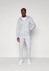 Sweatshirt cinzenta com fecho de correr, combinada com uma t-shirt branca e calças de fato de treino cinzentas, feitas de tecido macio. Apresenta punhos canelados e detalhes de marca.