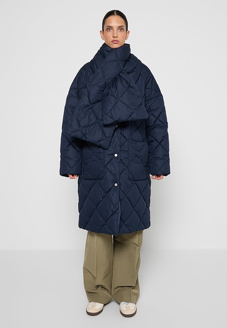 Granatowy pikowany płaszcz puchowy z dużym, opadającym kołnierzem, zatrzaskami oraz oversize'owym krojem. Stylizowany z szerokimi spodniami w kolorze khaki i obuwiem.