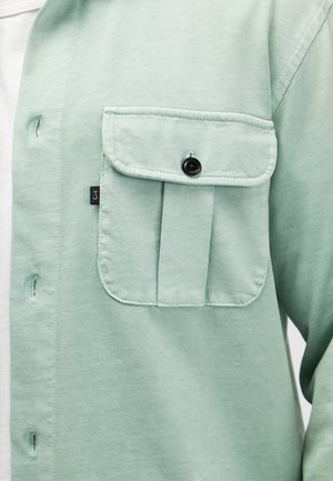 Lichtgroen overhemdzakje met een zwart geknoopt klepje en zichtbare stiksels op een kledingstuk met lange mouwen gedragen over een witte binnenlaag.