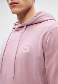 Roosa kapuutsiga pusakas, pehme tekstuuriga, rinnal on väikene ruudukujuline logo sobivas värvuses, ribakätised ja reguleeritav kapuuts.