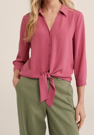 Femme portant un chemisier rose poussiéreux à manches longues boutonné et noué à la taille, avec un pantalon taille haute vert olive, main dans la poche, bague à la main droite.