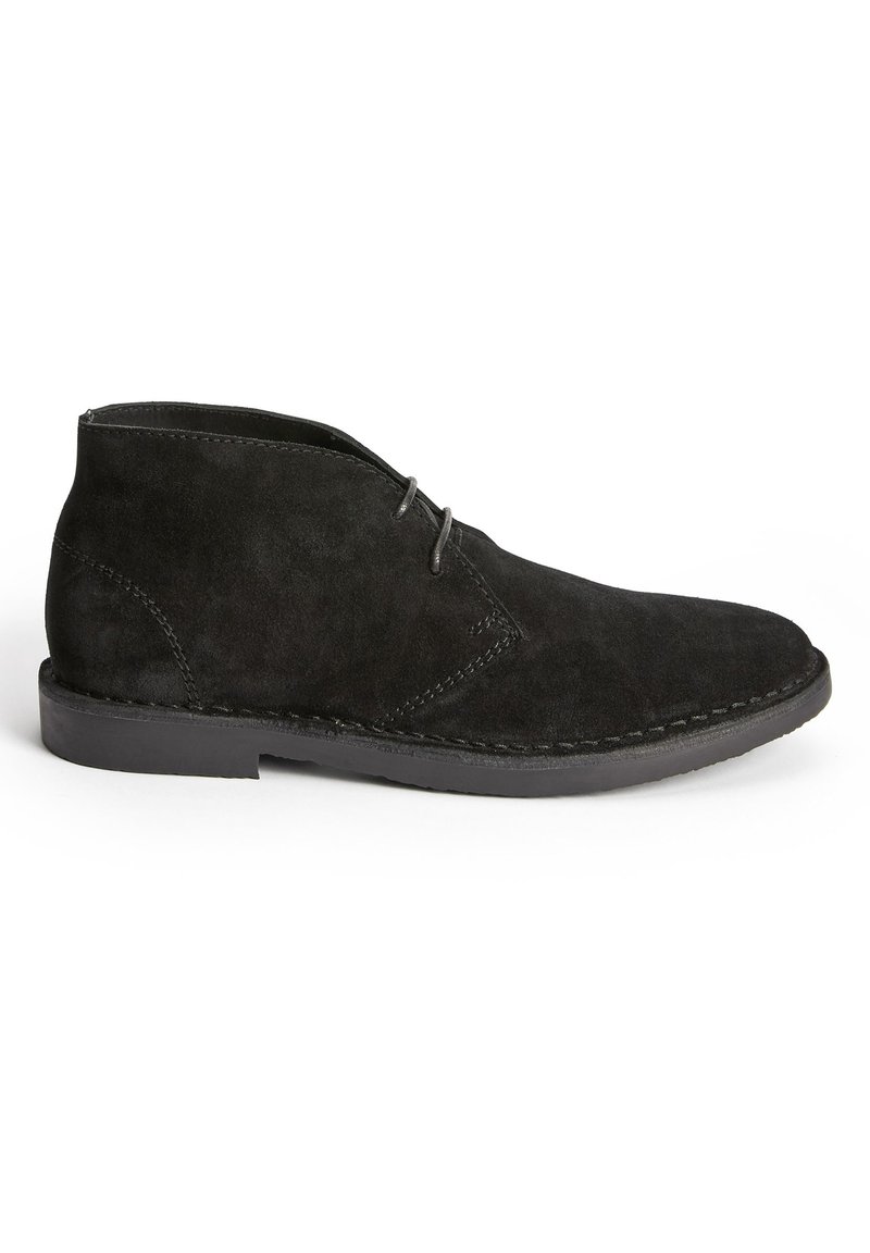 Botas de ante negras hasta el tobillo con punta redondeada, dos ojales para los cordones y una suela de goma plana con costuras visibles a lo largo de los bordes.