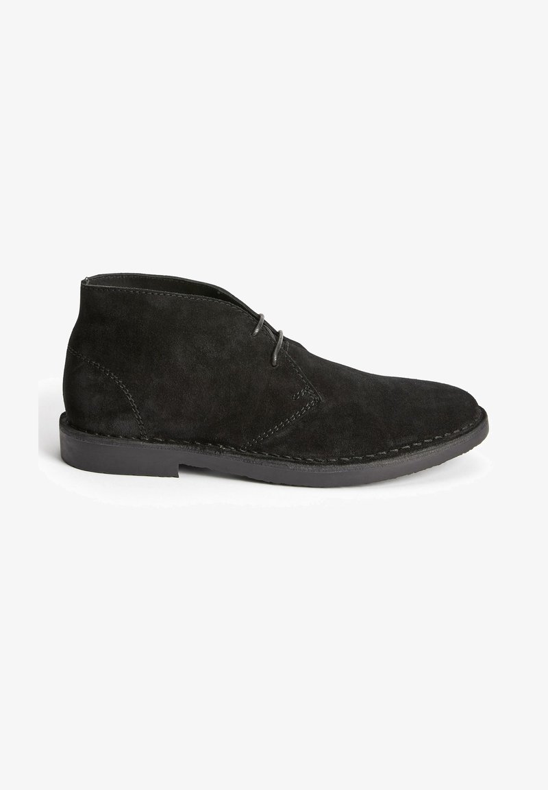 Bottines en daim noir avec un bout arrondi, deux œillets pour les lacets et une semelle en caoutchouc plate présentant des coutures visibles le long des bords.