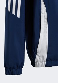 Veste marine avec des accents blancs, présentant une texture lisse, des poignets élastiques et trois rayures blanches sur l'épaule. Poche latérale visible.