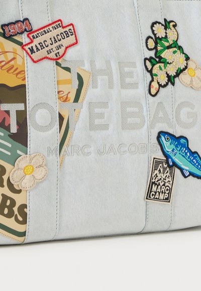 Sac cabas en denim clair avec des écussons brodés de fleurs, un poisson bleu, des badges avec du texte et des motifs sur le thème de l’aventure. Texte visible : "THE TOTE BAG".